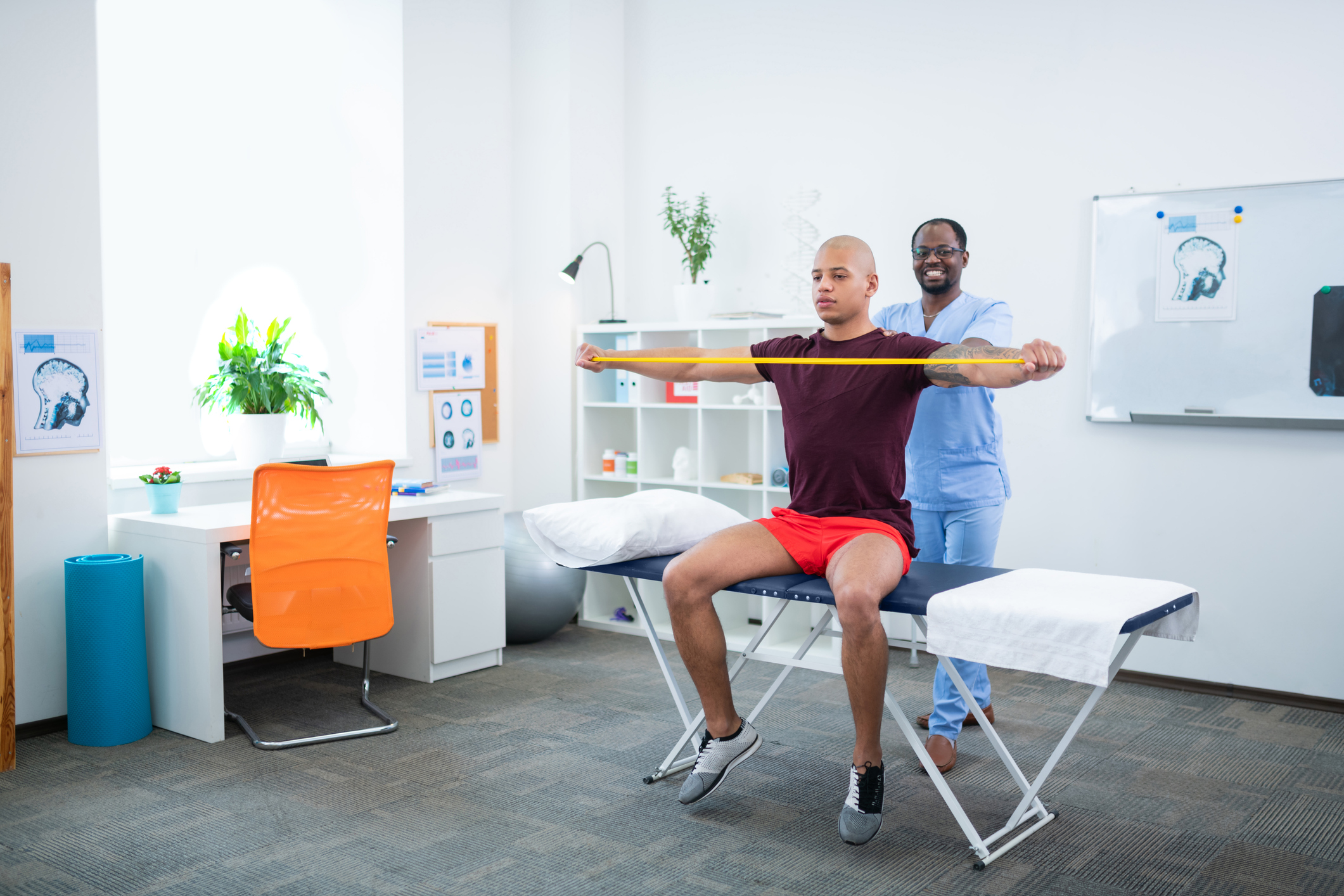 Smiling therapist. Dark-skinned sports therapist smiling while stretching arms of sportsman