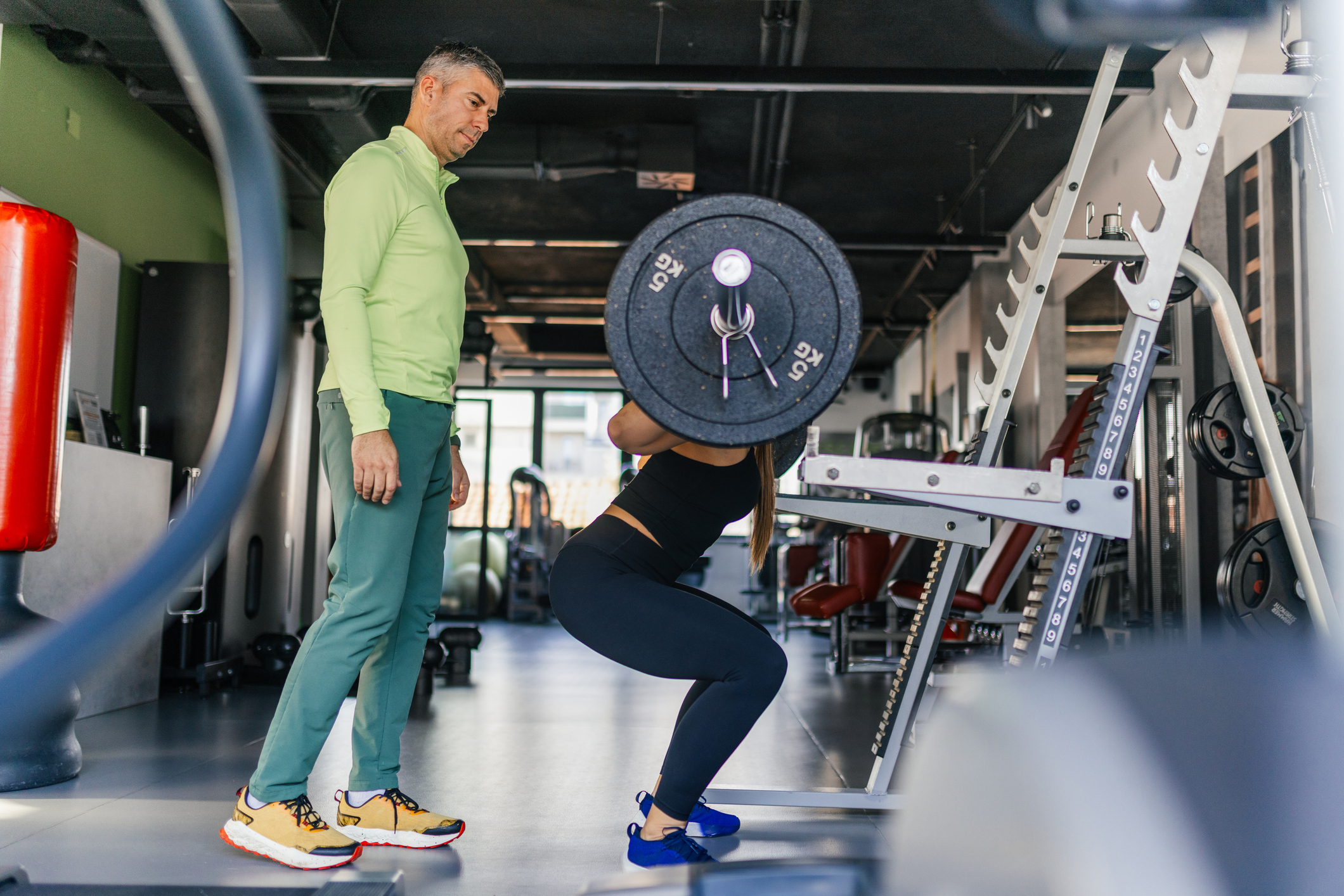 A strong, athletic woman doing squats with weights in modern gym, while her trainer assist and helping her the proper technique. She is dressed in sporty workout clothes