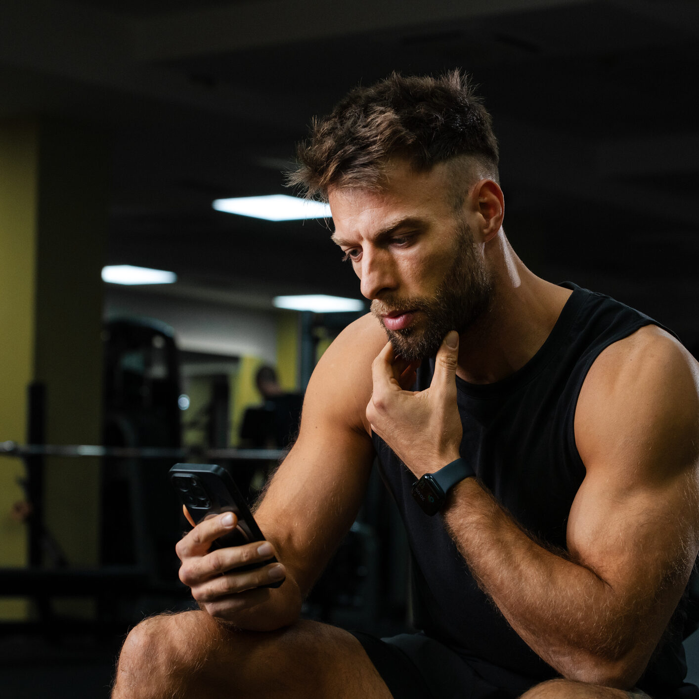 A muscular man intently watches his smartphone screen in a modern gym, embodying the rise of digital fitness. His posture and expression convey concentration  great for tech brands, wellness blogs, and remote training platforms.