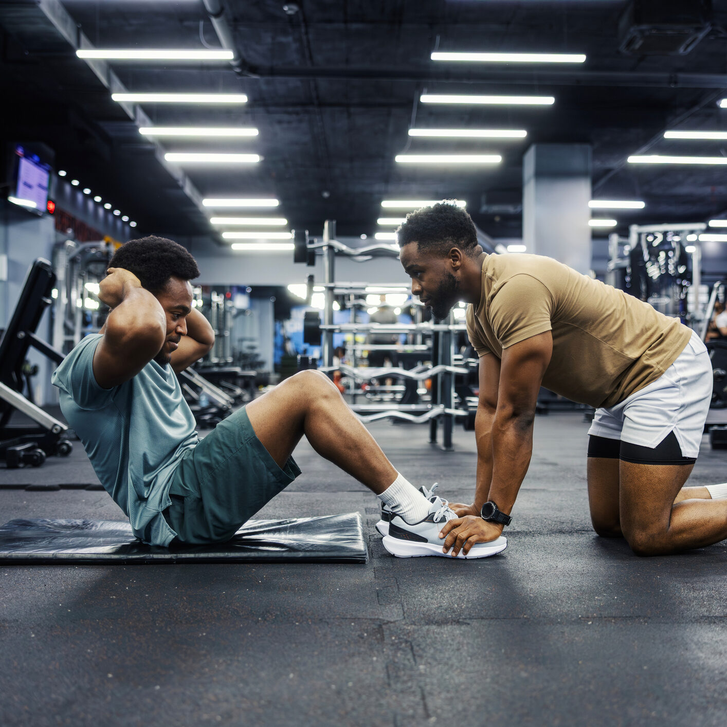 Male personal trainer supporting a man's feet during sit-ups on an exercise mat, focusing on core strength